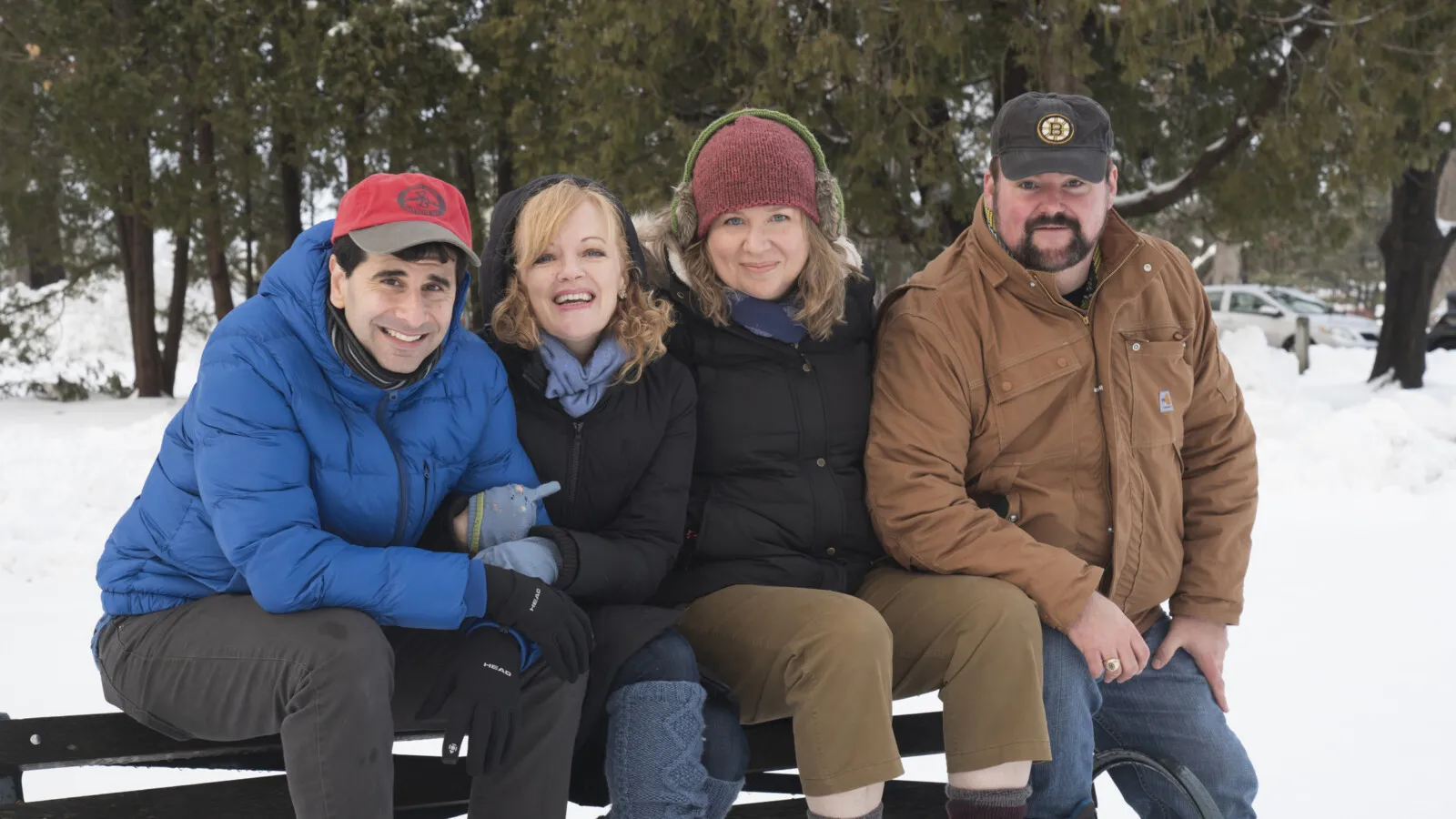 The image shows a group of four posing together on a bench in the snow, with one man wearing a brown jacket and sunglasses, a woman donning a red hat, and the others both bundled up in winter clothes. They are all smiling and looking at the camera, creating a warm and happy atmosphere despite the cold weather.