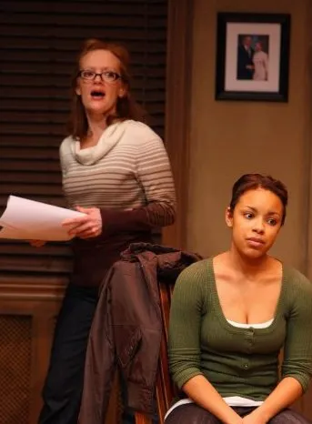 Two women are present, one is standing and the other is sitting. The woman standing has her mouth wide open while the woman sitting looks at something off to the side of the camera.
A person reading a piece of paper in front of themself.
The woman with her mouth open appears surprised or shocked.
The woman sitting on a chair with her hands folded next to her.