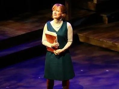A woman stands on a stage, dressed in a green dress and holding an orange book. She is smiling as she looks to the side. The stage has wooden floors and there are stairs behind her. There is also some text that says "1940's".
