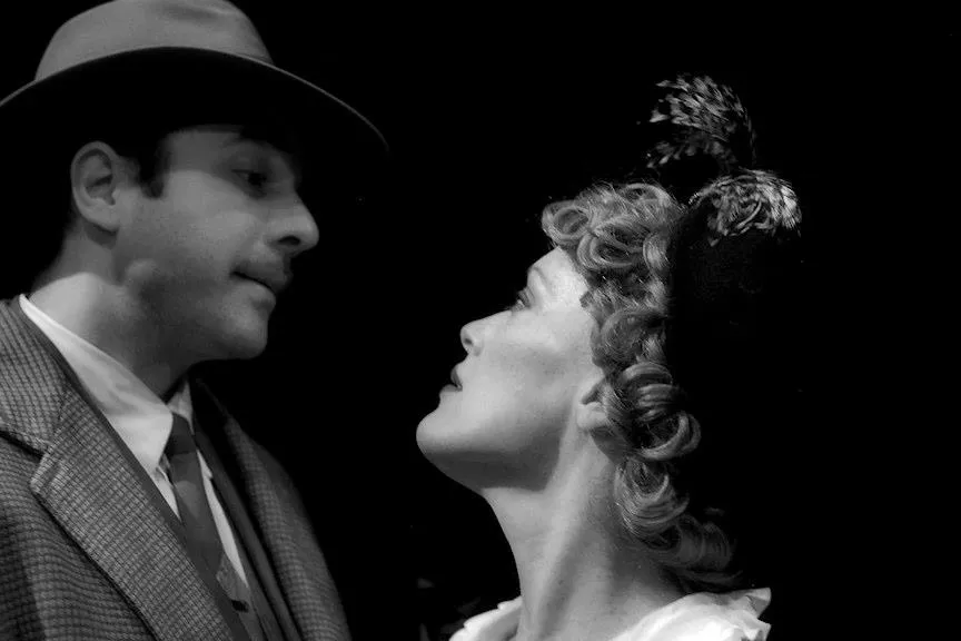 A man standing next to a woman with a flower on her head. The woman is looking upward toward the sky. They are both wearing hats. The picture has a black and white tint.