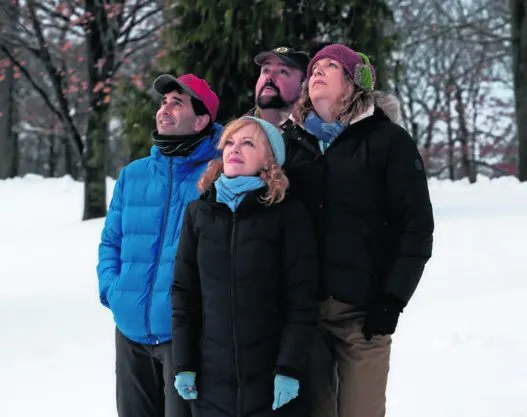 A group of four is standing outside, bundled up against the cold weather with hats on their heads. The two adults are wearing jackets, one blue and the other black, while the others are sporting a blue coat and a pink hat respectively. They all have rosy cheeks, suggesting they've been out in the chilly air for a while. Behind them, a snow-covered landscape stretches out under a clear sky.
