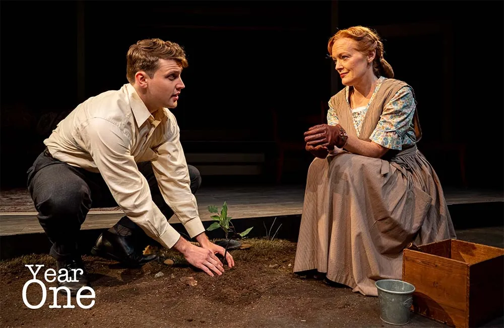 A woman is sitting on a wooden bench outside with a man kneeling down next to her looking at the ground. The woman has a basket of food beside her. A tree trunk with roots jutting out from it can be seen near them, as well as a bird and some plants in front of them. In the background there is a small stage with chairs on it. The text "Year One" appears in the bottom left corner of the image.