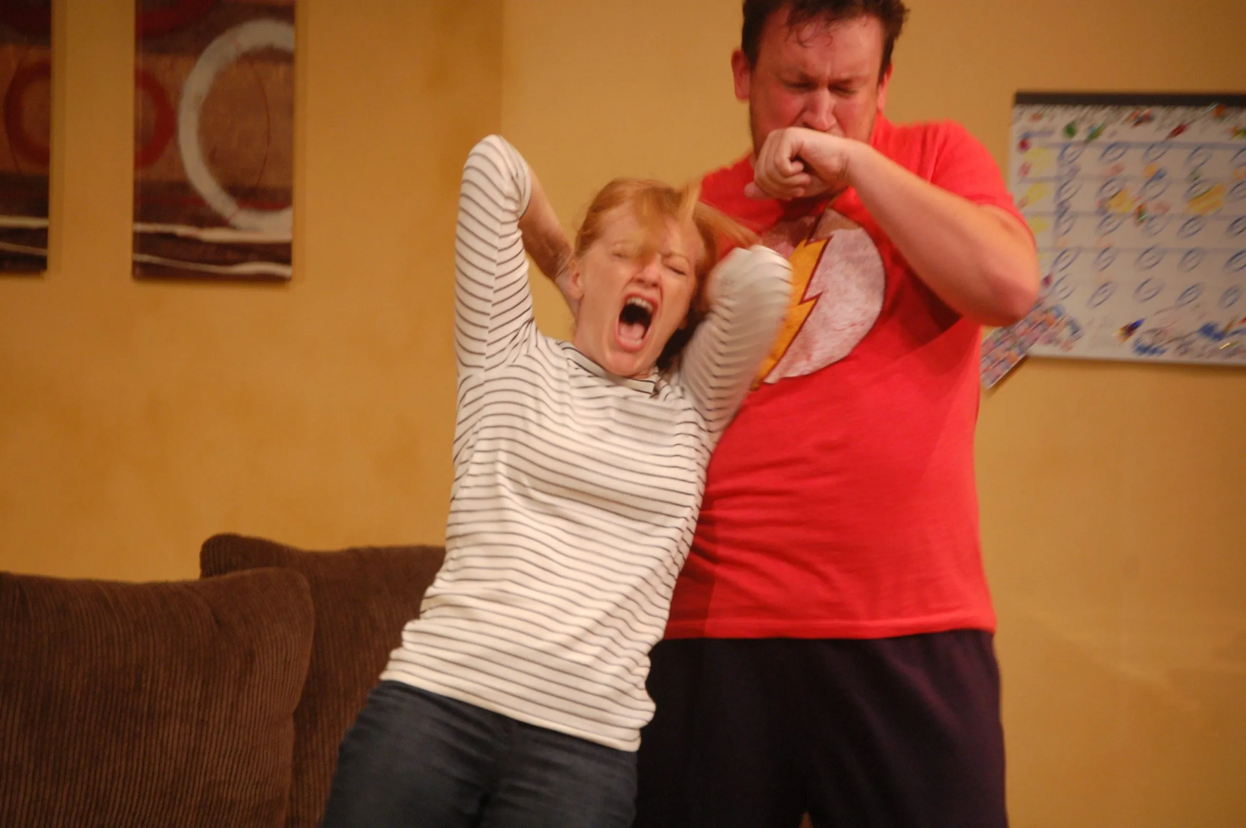 In the image, a man wearing a red shirt is standing behind a woman with her arms raised. The woman has a surprised expression on her face. The setting appears to be indoors as there are couches in the background along with pictures hanging on the wall. There's also a calendar present that suggests this might have been taken during the month of December.