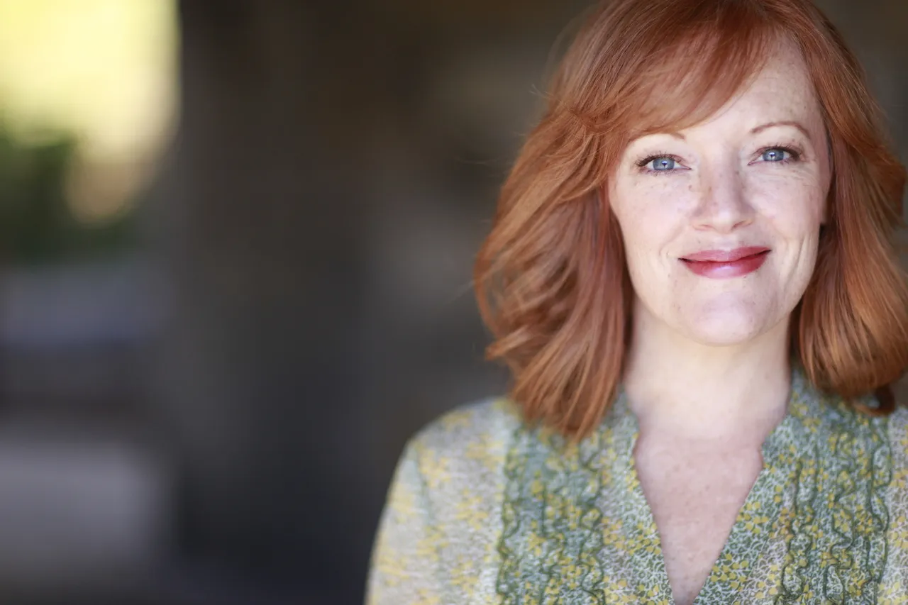 A woman with red hair is wearing a patterned blouse. She has blue eyes and is smiling at the camera. The background is out of focus, but it appears to be an urban setting with buildings in the distance.