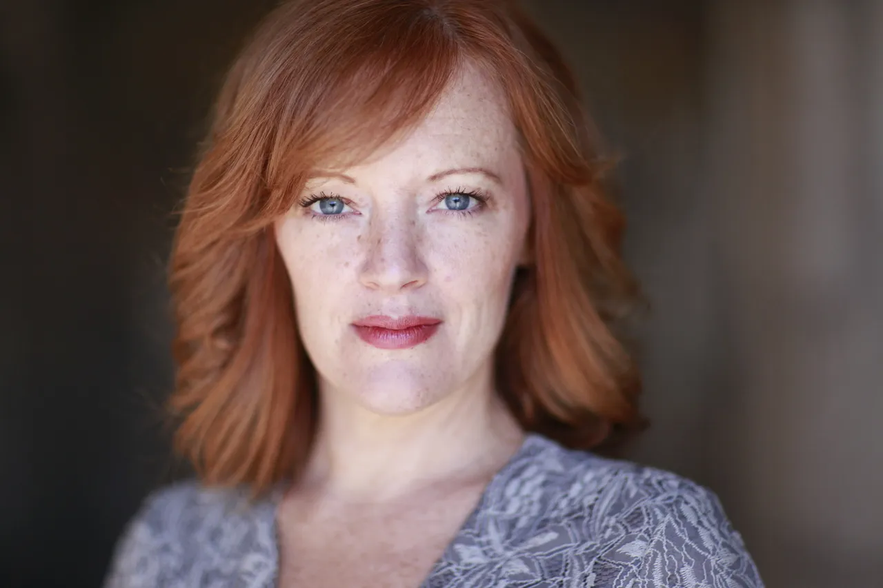 A woman with reddish blonde hair wearing a gray top is looking at the camera.