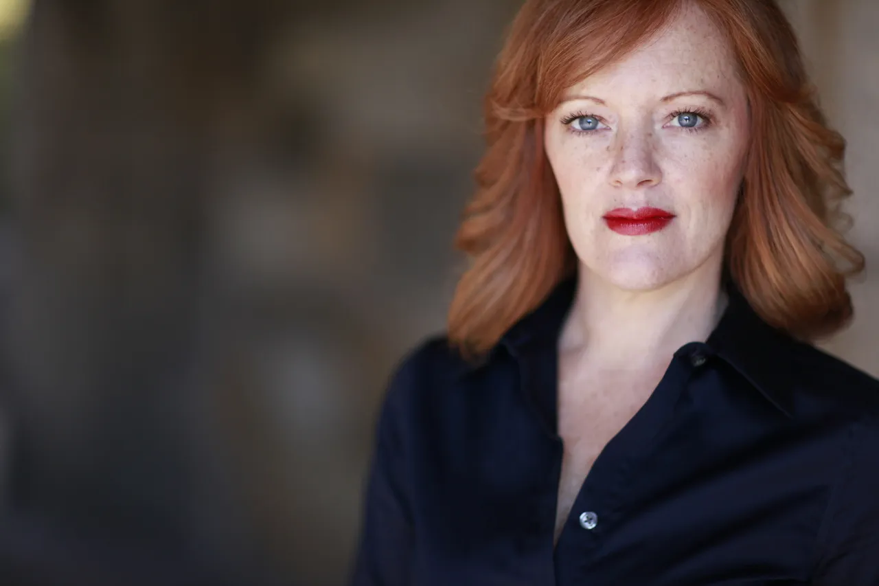 The woman is wearing a black blouse with ruffles around the neckline and has red lipstick on her lips.