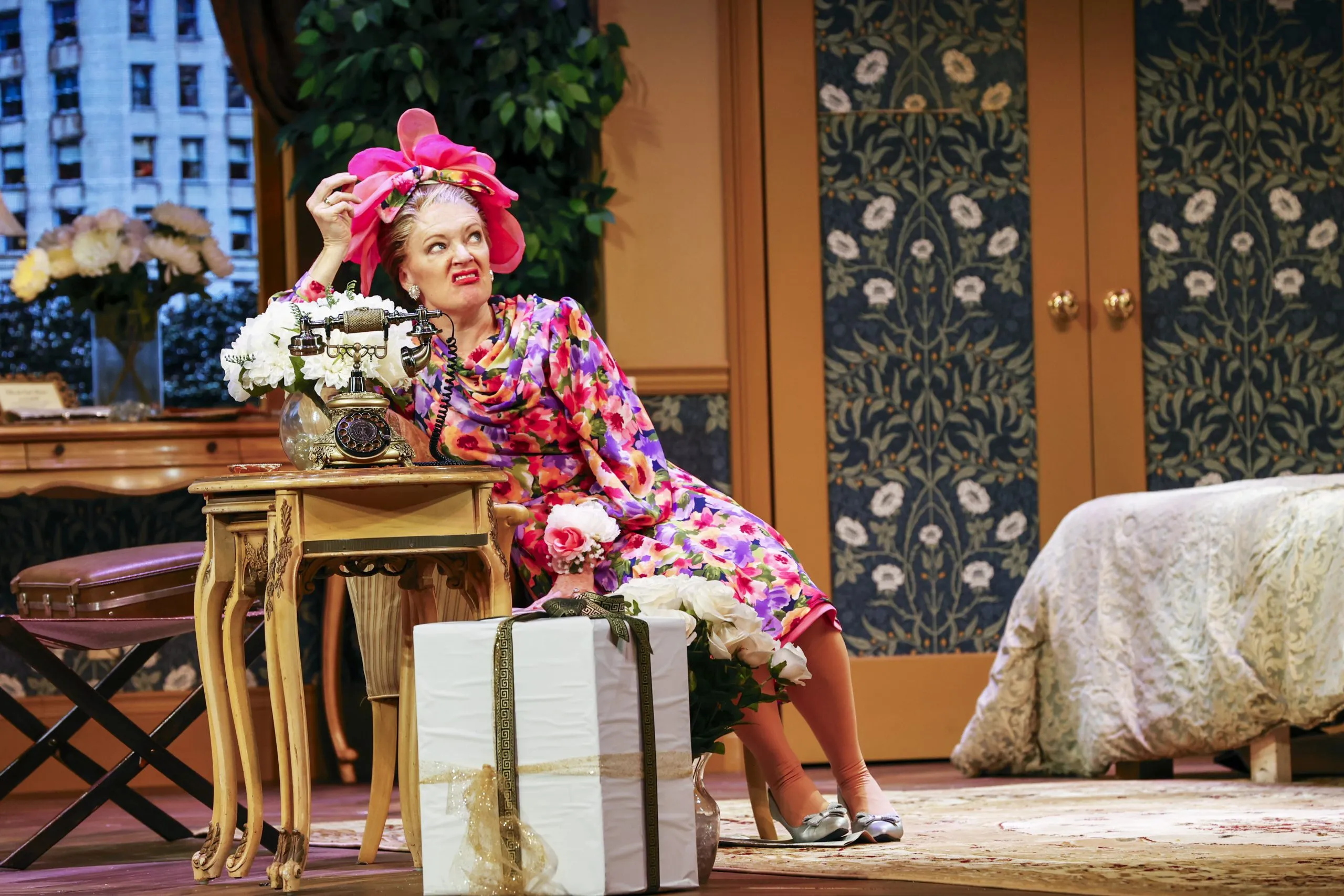 A woman sitting on a table with her hand on her head and flowers on the table.
The room has a floral patterned wallpaper, a brown door and some furniture including two benches.