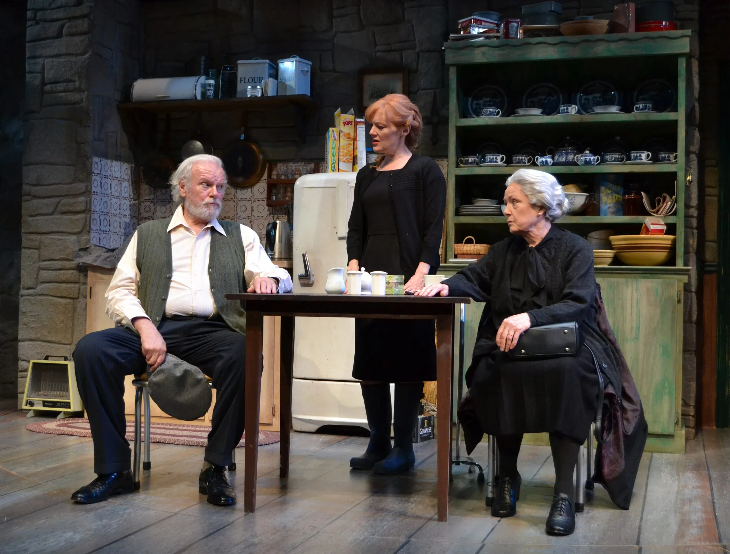 The man is sitting down, the woman is standing up next to him and a lady with a black coat is also there. There is an old fridge in their house that contains bowls on it.
It seems like this couple is enjoying tea together while being surrounded by pots and pans.