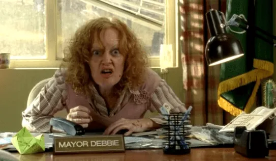 Mayor Debbie is sitting at the office desk with her arms crossed.