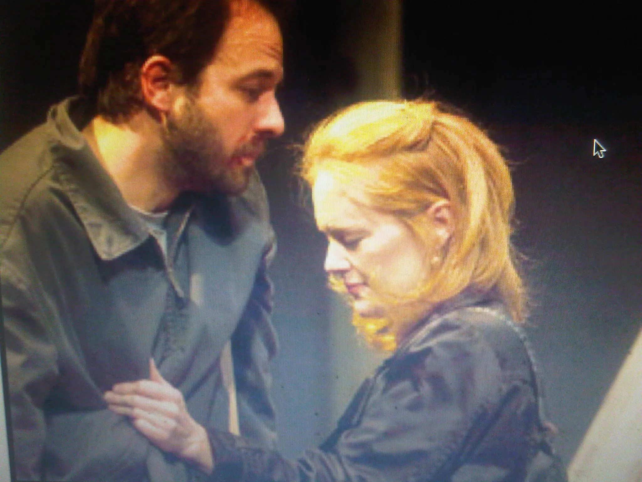 A man stands behind a woman with blonde hair, both are looking downwards. She cries.