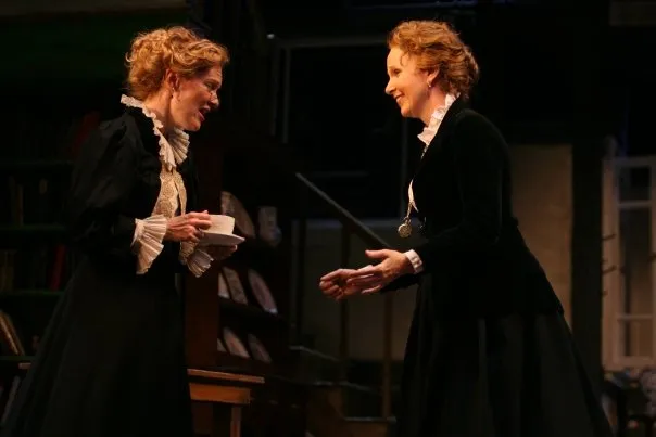 Two women, both dressed up and holding a cup of coffee or tea, are standing next to each other. They appear to be engaged in conversation as they hold their drinks close to them. The woman on the left is also wearing glasses, adding an intellectual touch to her appearance. They seem to be in a well-lit room with a bookshelf visible behind them, suggesting that they might be in a library or a similar setting. Their exact location isn't clear from the image, but their expressions and attire suggest a casual yet sophisticated atmosphere.