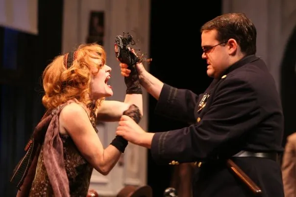 On stage, a man dressed as a soldier hands over his hat to an actress who is crying. The actor has a microphone in front of him while the actress holds up her hand with the hat. The background features a blue curtain and white columns.