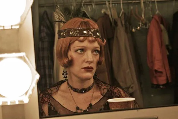 The woman is wearing a large black feathered headdress with tinsel trim. She has red colored hair, and her face bears a serious expression.
She is holding a white cup in her hand and looking at it. Her reflection can be seen in the mirror behind her.