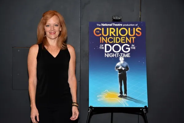 A red haired woman with a black shirt on is standing next to a poster of the movie "Curious Incident". The title of the movie is shown at the top of the poster, and there's an image of a man in glasses on it too. The poster is displayed on a black stand.