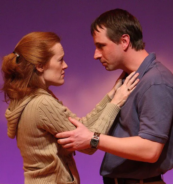 A woman with red hair wearing a beige sweater is embracing a man who has a watch on his wrist. They are standing against a purple background.